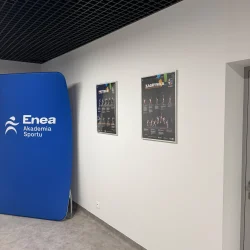 Niebieski baner Enea Akademia Sportu i plakaty instruktażowe na białej ścianie korytarza.