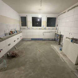 Pomieszczenie w trakcie remontu z surową posadzką i częściowo ułożonymi białymi płytkami na ścianach.