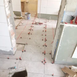 Układanie jasnych płytek podłogowych z użyciem systemu poziomowania w remontowanym wnętrzu.