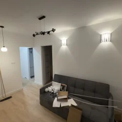 Jasny salon po remoncie z szarą sofą, na której leżą kartony i elementy montowanego oświetlenia.