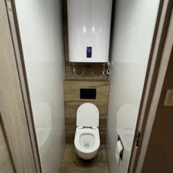 Wąska toaleta z podwieszanym WC i dużym bojlerem, wykończona białymi i drewnopodobnymi płytkami.