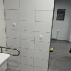 Ściana wyłożona białymi płytkami z włącznikami światła i widok z łazienki na sąsiednie pomieszczenie.